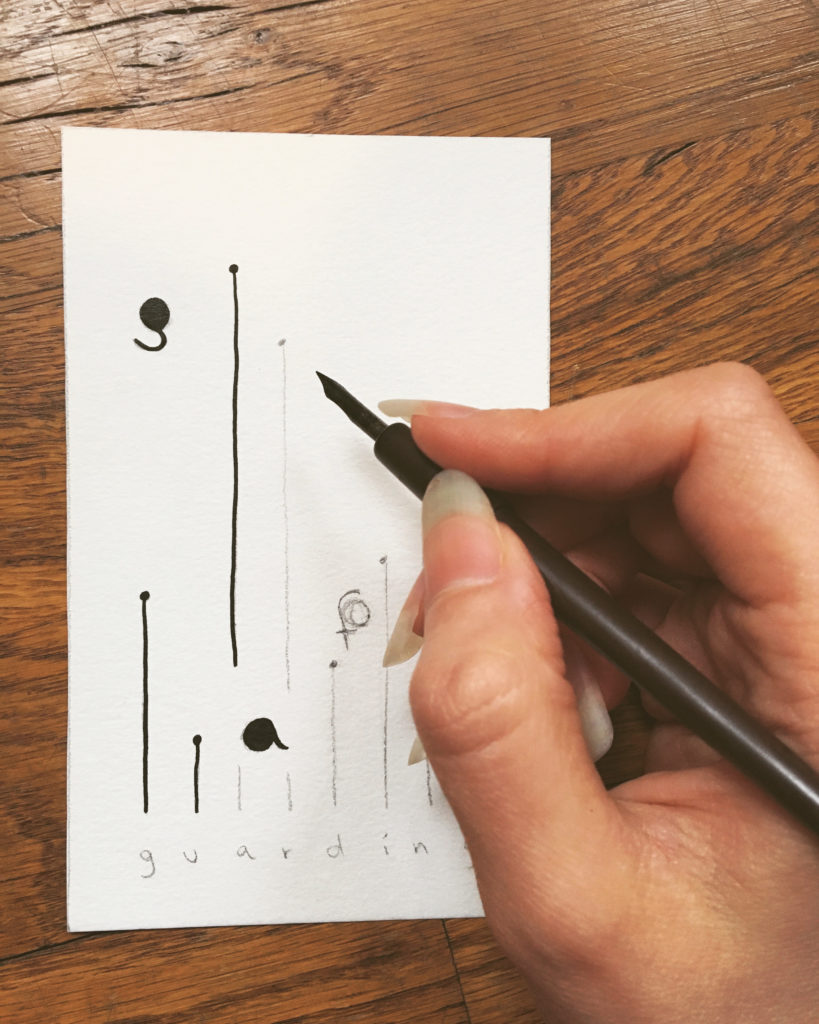 Calligraphy pen in hand, Alexa Dexa traces over a pencil design on 300gsm sketchbook paper using black ink. The design features the text “safe guarding” with the letters of “safe” sprawled out throughout the height of the card, each letter shaped into a large circle. “gaurding” is written across the bottom of the card, each letter sprouting an upwards straight line of varying lengths attaching to its own small circle.